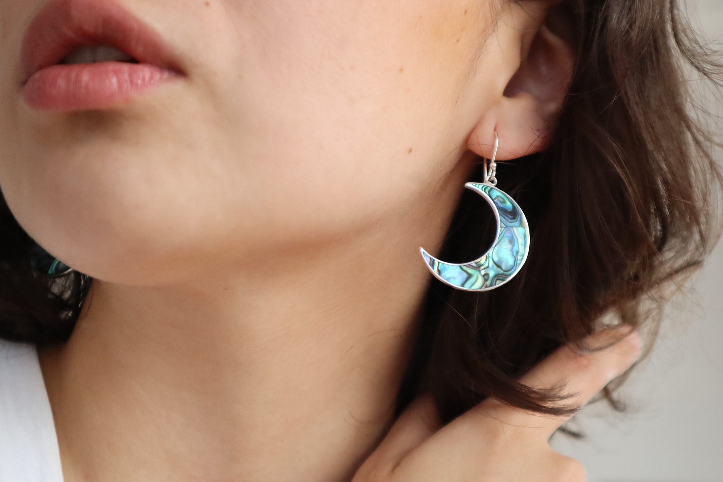 Close-up of a person wearing a green blue crescent moon earring made of abalone shell